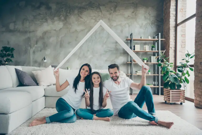 Famille souriante assise sur un tapis dans un salon, formant un toit symbolique au-dessus de leur tête avec deux baguettes blanches.