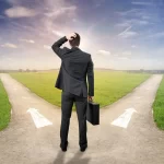 Homme en costume hésitant devant un chemin qui se divise en deux directions sous un ciel dégagé.