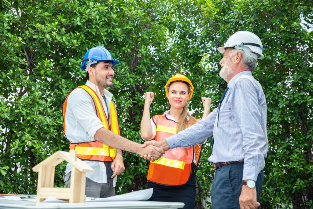 Un courtier immobilier serre la main d’un inspecteur en bâtiment portant un casque, tandis qu’une acheteuse sourit à côté, devant une maison.