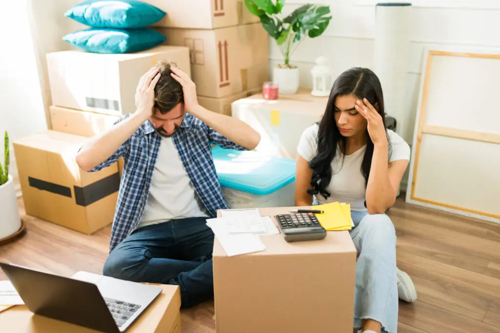 Un couple stressé assis sur le sol entouré de cartons, regardant des documents et un ordinateur avec frustration, illustrant les défis d’un achat immobilier sans accompagnement humain.