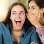Deux jeunes femmes discutent en secret, l’une chuchotant à l’oreille de l’autre, illustrant la dissimulation d’un conflit d’intérêts en immobilier.