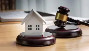 Maison miniature posée sur un socle à côté d’un maillet de juge, illustrant le cadre légal et la protection en courtage immobilier.