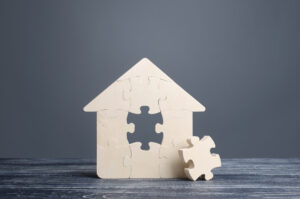 Maison en bois en forme de casse-tête avec une pièce manquante posée à côté, illustrant qu’il manque un élément essentiel à l’acheteur(euse) sans véritable représentation titre Traitement équitable en immobilier et pièce manquante de la représentation légende Une maison incomplète pour illustrer qu’un traitement équitable ne remplace pas la vraie représentation de l’acheteur(euse) description Image d’une maison en bois composée de pièces de casse-tê