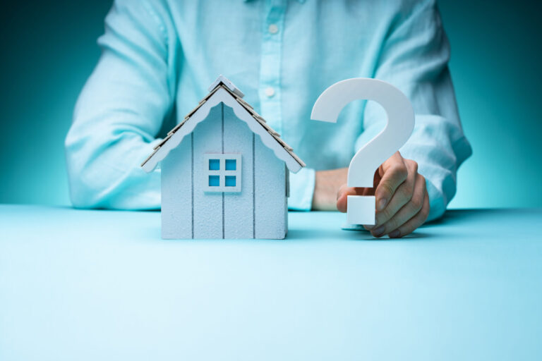 Main tenant une maquette de maison blanche avec un point d'interrogation bleu au-dessus, symbolisant les questions sur l'achat immobilier.
